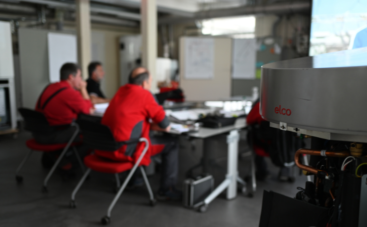 Personen in rode werkjassen tijdens een vergadering in een technische ruimte, op de voorgrond een radar- of meetapparaat.